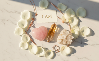 Luxury flat lay of affirmation crystals including rose quartz heart, citrine and clear quartz on white marble with white rose petals and gold I AM card