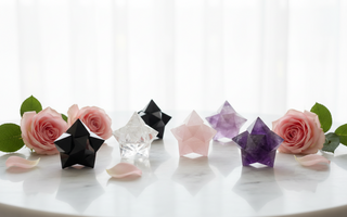 A collection of Merkabah star tetrahedron crystals in black obsidian, clear quartz, rose quartz and amethyst arranged on a white marble surface with soft pink roses
