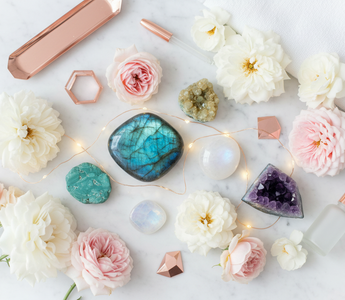 Luxury flat lay of new beginning crystals including labradorite, rainbow moonstone, chrysocolla, citrine and amethyst with blush roses and fairy lights on white marble