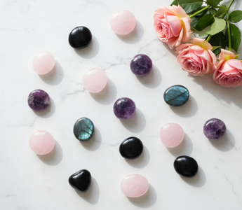 Assorted crystal palm stones and worry stones including rose quartz, amethyst, black obsidian and labradorite arranged on white marble with soft pink roses