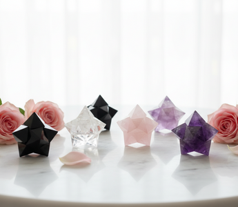 A collection of Merkabah star tetrahedron crystals in black obsidian, clear quartz, rose quartz and amethyst arranged on a white marble surface with soft pink roses